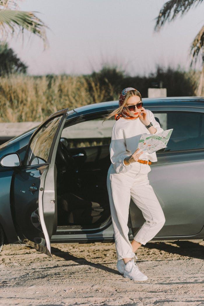 A fashionable tourist stands by an open car door, reading a map under sunny skies.