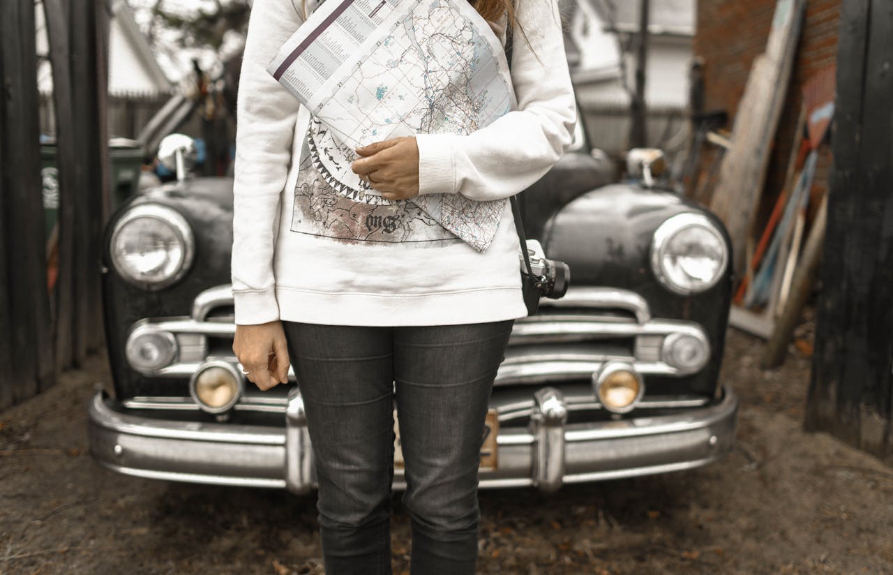 Woman holding a map and camera in front of a vintage car, ready for an adventure.